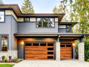 Front view of a contemporary two-story house with wood-paneled double garage doors, stone columns, large windows, and lush landscaping.