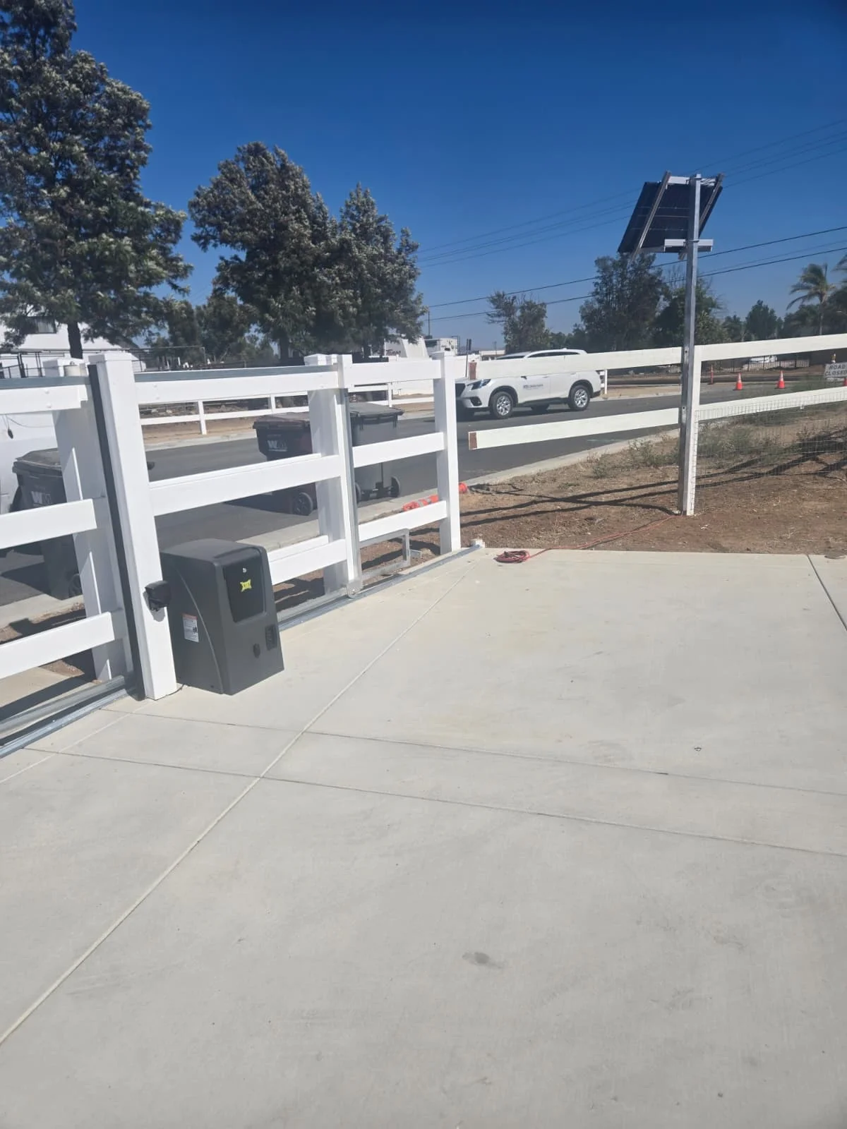 Automatic sliding gate installed with solar-powered system and white vinyl fencing on a concrete driveway.